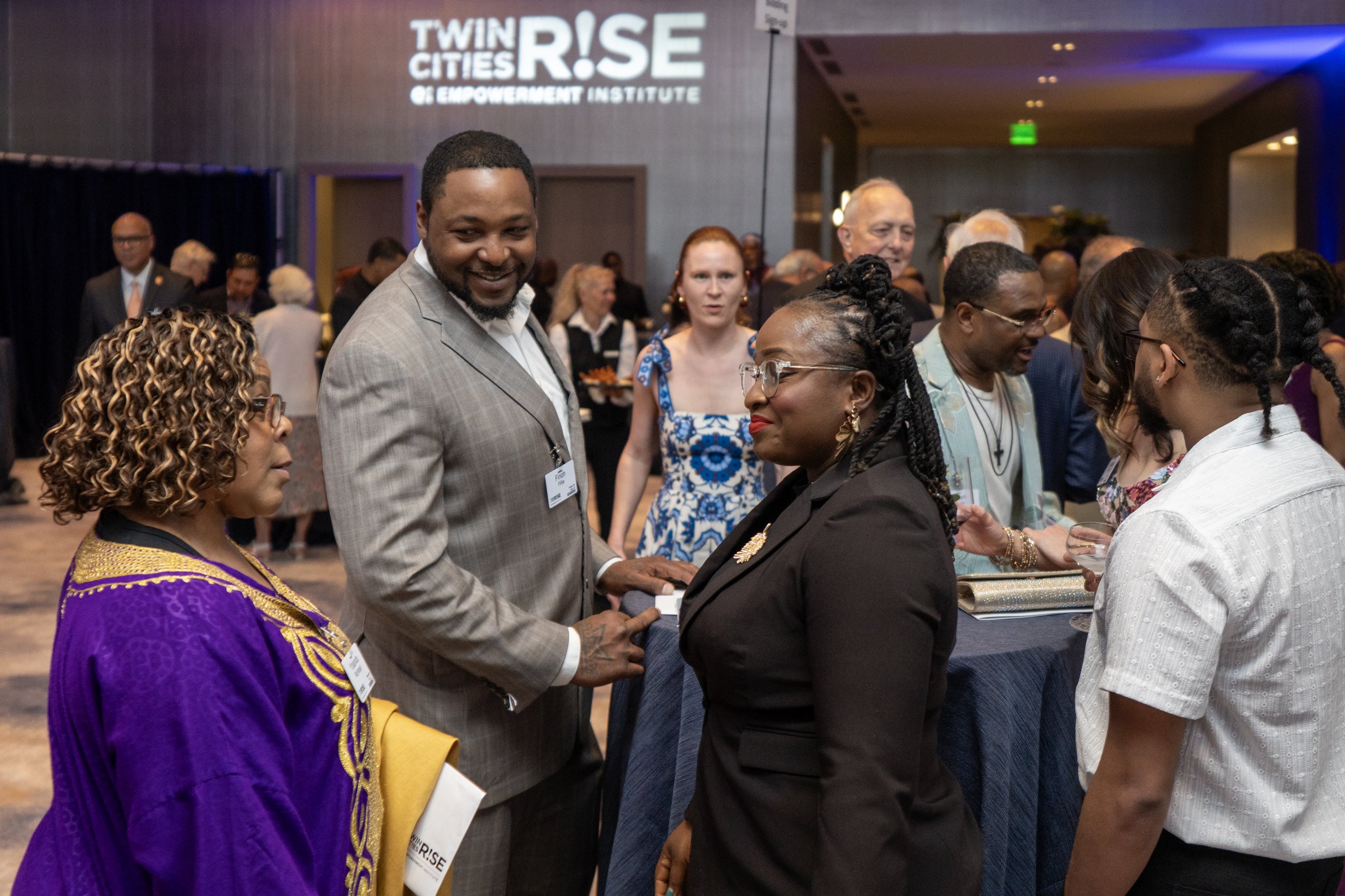 Group of diverse people at an event, engaging in conversation, with a sign that reads 'Twin Cities Rise' in the background.