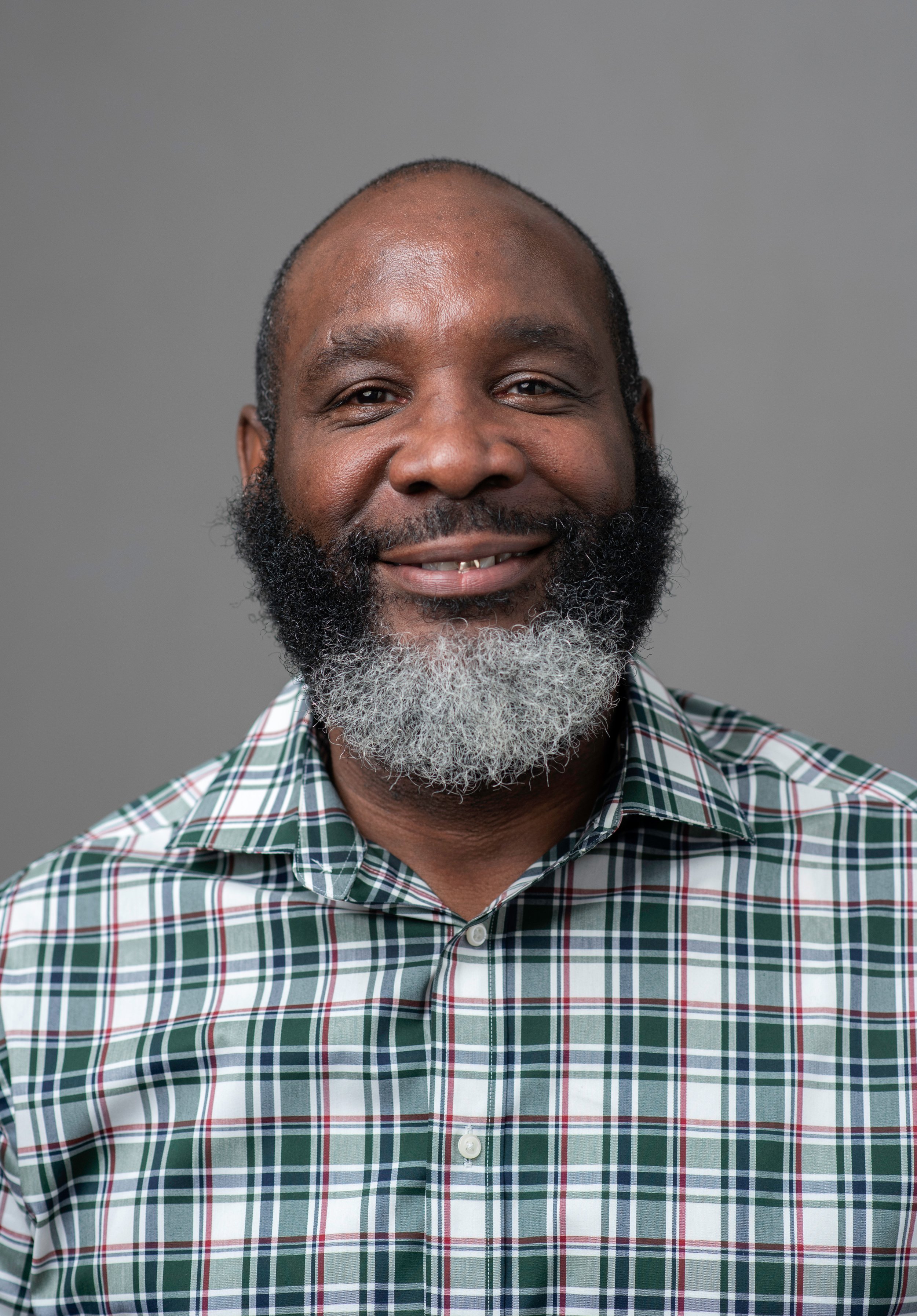 Close-up of a middle-aged African American man with a beard, wearing a green, red, and white plaid shirt, smiling against a plain gray background.