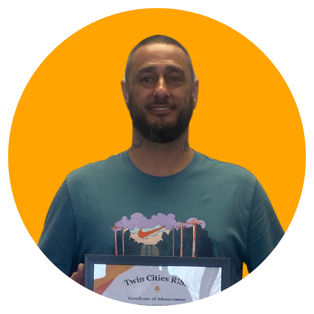 A man with a beard smiling and holding a framed certificate, standing in front of a bright orange background.