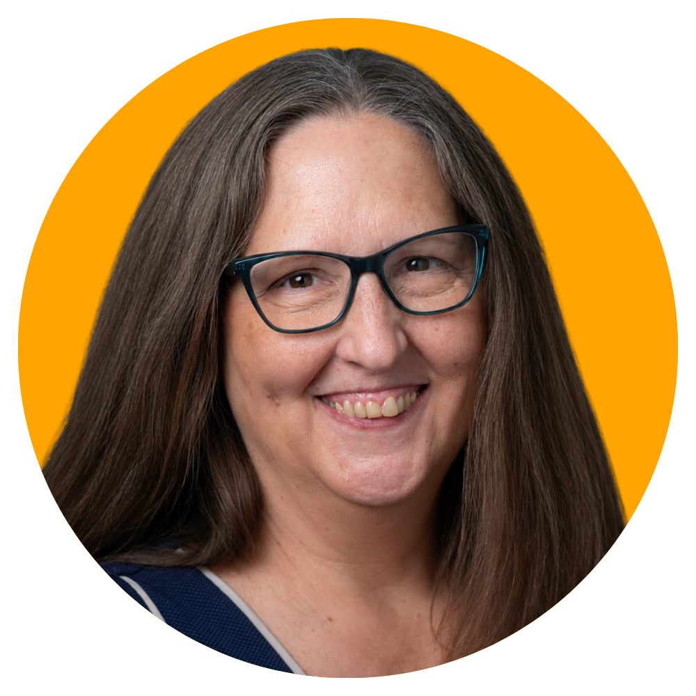 A woman with long brown hair, glasses, and a smile, wearing a dark shirt with white trim, against a yellow background.