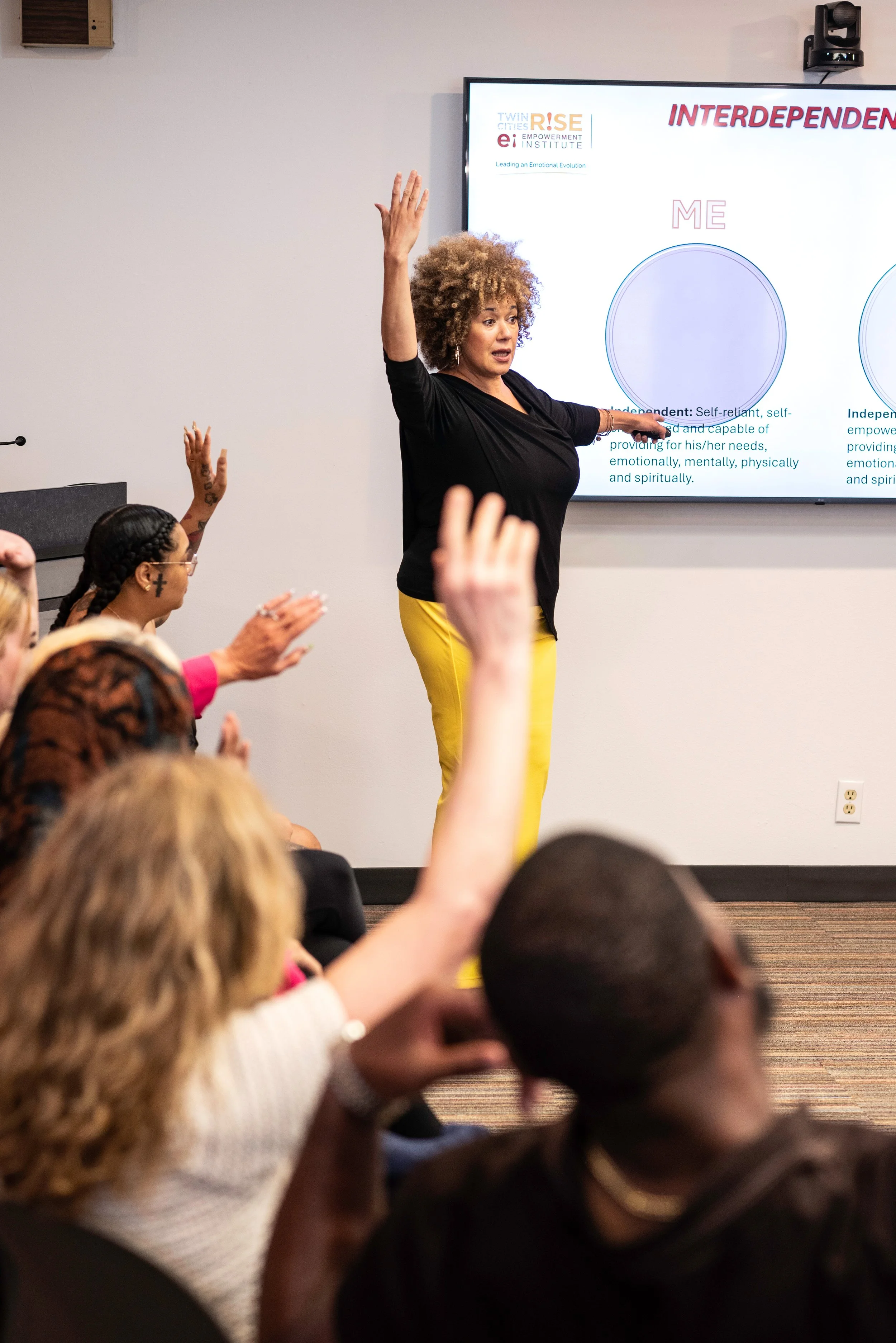 A woman with curly hair giving a presentation in front of a screen to a diverse group of people. The screen displays a diagram with the words "INTERDEPENDENCE" and "ME".