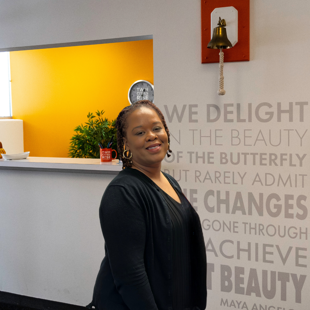 A woman with dark hair and earrings smiling in an office lobby area. Behind her is a wall with a quote about beauty and change, and a wall clock. There's a reception desk with a plant and a mug.