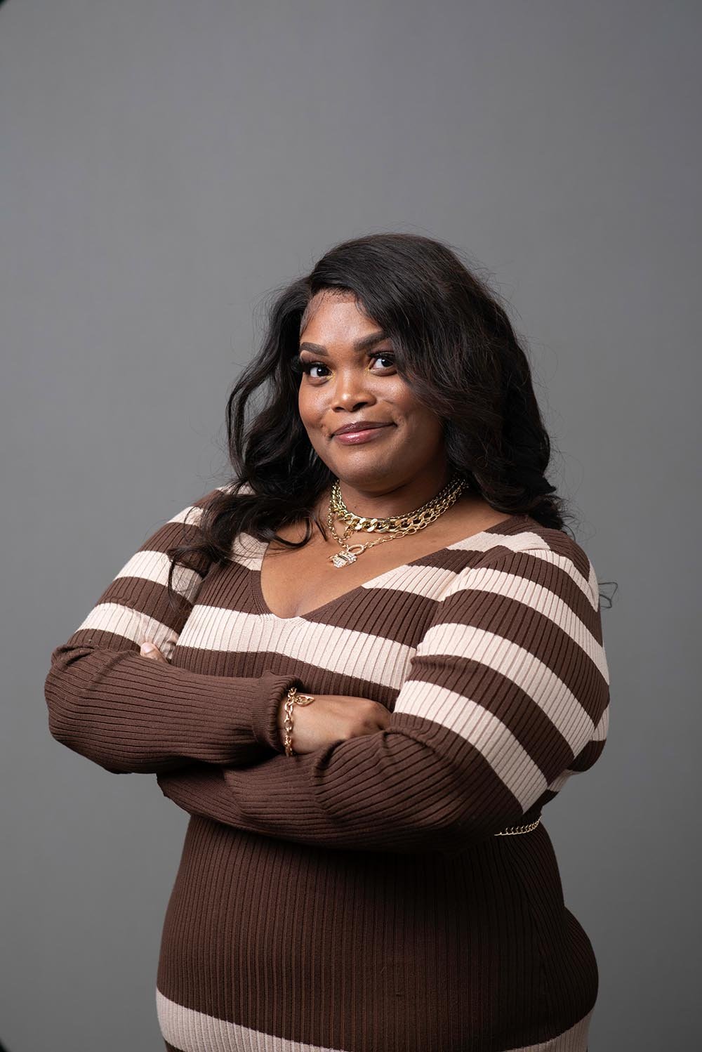 A woman with dark hair, wearing a brown and beige striped sweater, gold chains, and jewelry, standing with arms crossed against a gray background.