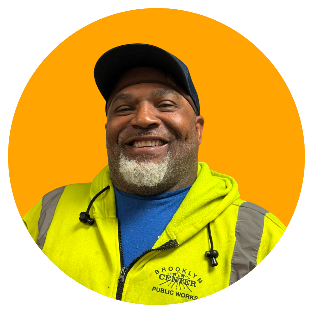 Smiling man wearing a yellow safety vest and a blue cap, with a beard, against an orange background.