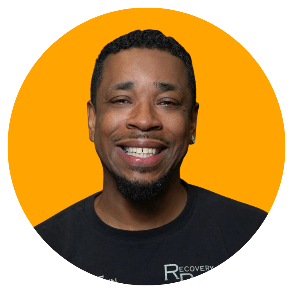 A smiling man with short curly hair, a goatee, wearing earrings, dressed in a black t-shirt, against a solid orange background.