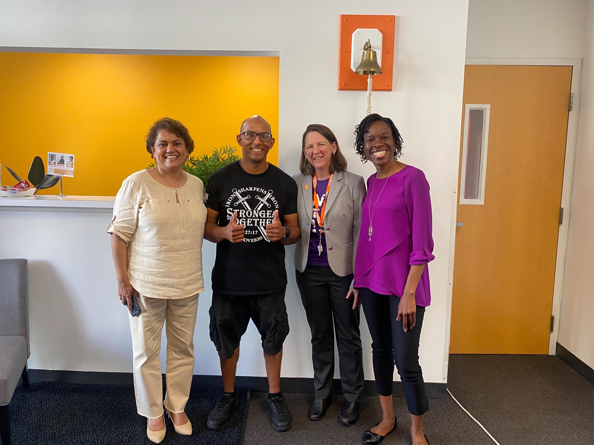 Three women and one man standing together in an indoor setting, smiling at the camera. The man in the center is wearing a black t-shirt with white text and giving a thumbs-up. There is a yellow wall behind them and a door on the right side.