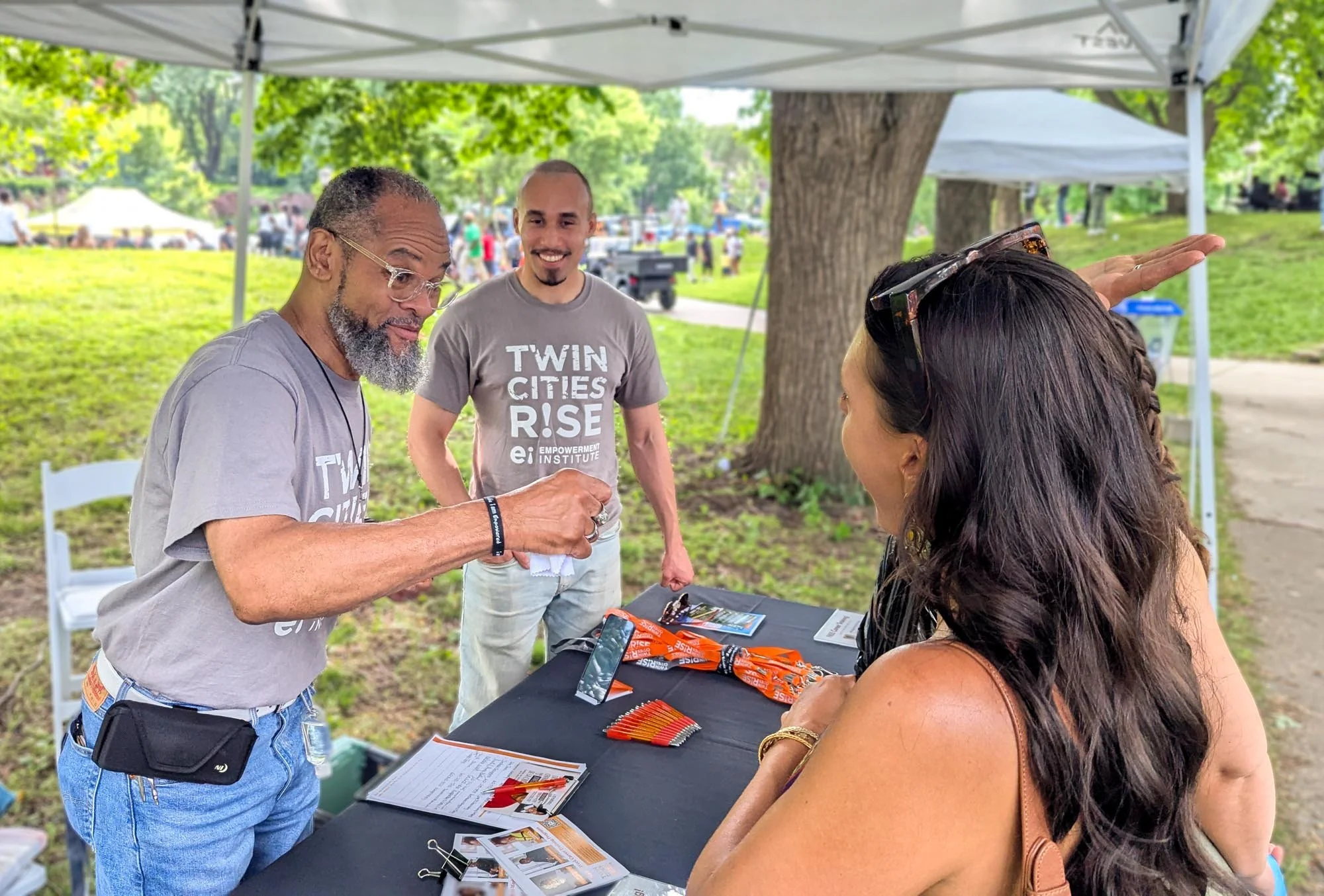 Three people at an outdoor booth under a canopy, engaging in a conversation. The booth has red, orange, and white promotional materials on the table. One person is handing an item to a woman, who is wearing sunglasses on her head. In the background, people and trees are visible.