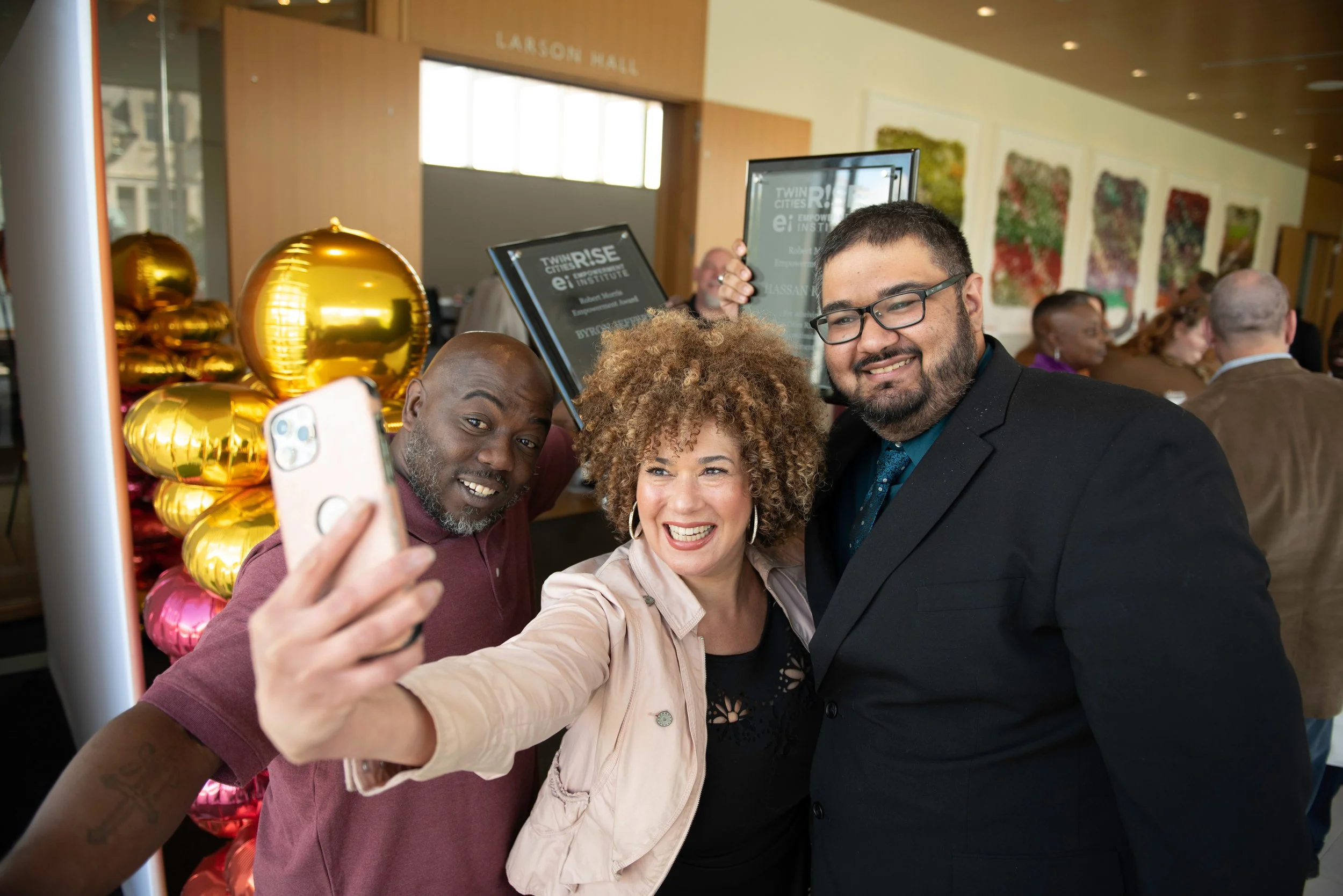 Three diverse people taking a selfie together at a formal event in a decorated venue with colorful balloon arrangements.