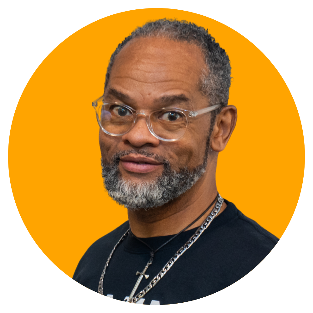 A middle-aged man with gray hair and a beard, wearing clear glasses and jewelry, looking at the camera with a slight smile. Background is orange.