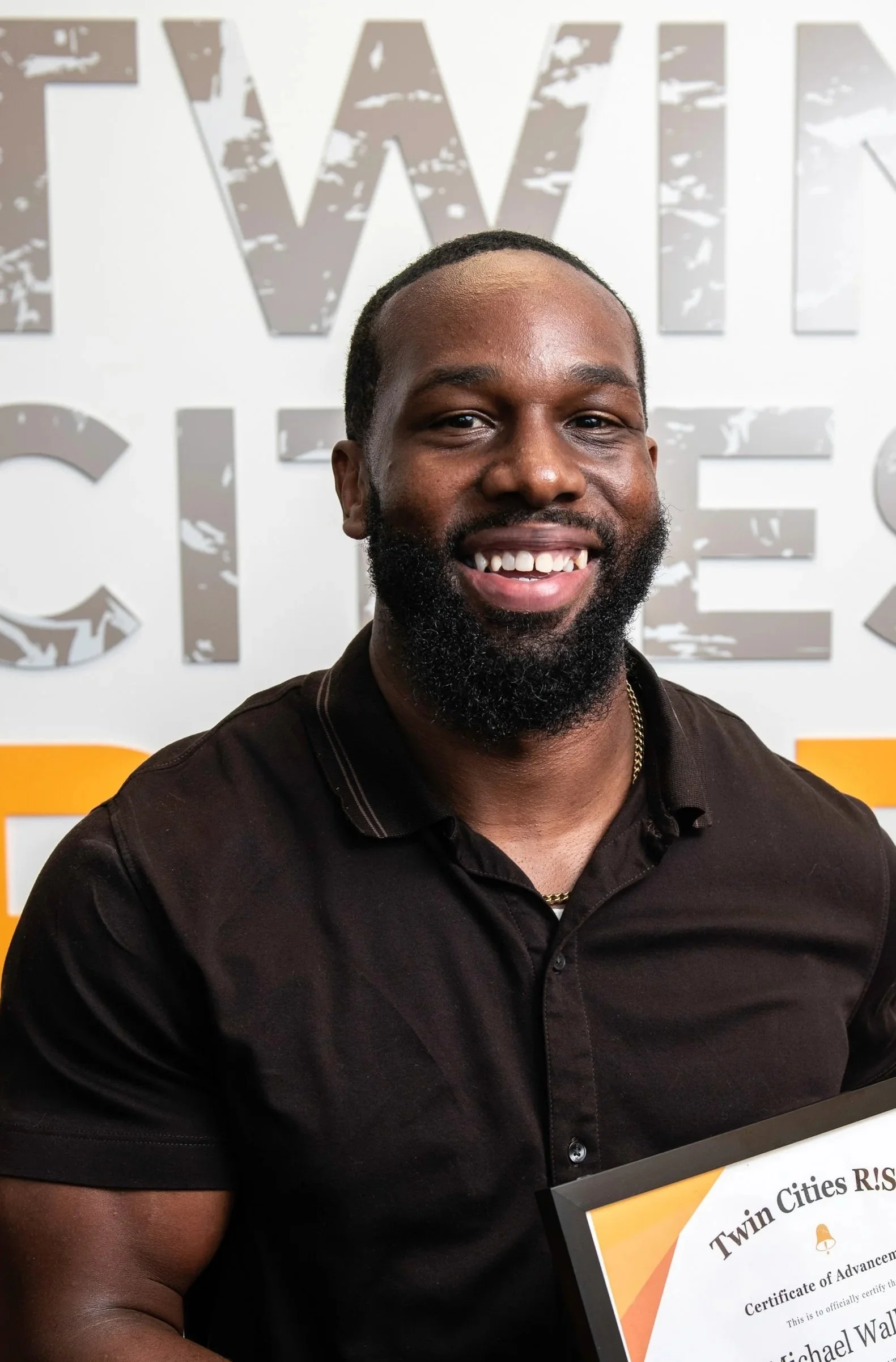 A smiling man with a beard holding a certificate of achievement in front of a wall with the Twin Cities RISE logo.