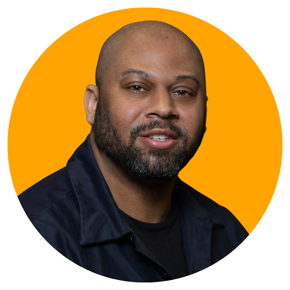 Close-up portrait of a man with a bald head and beard, wearing a dark jacket, against an orange background.
