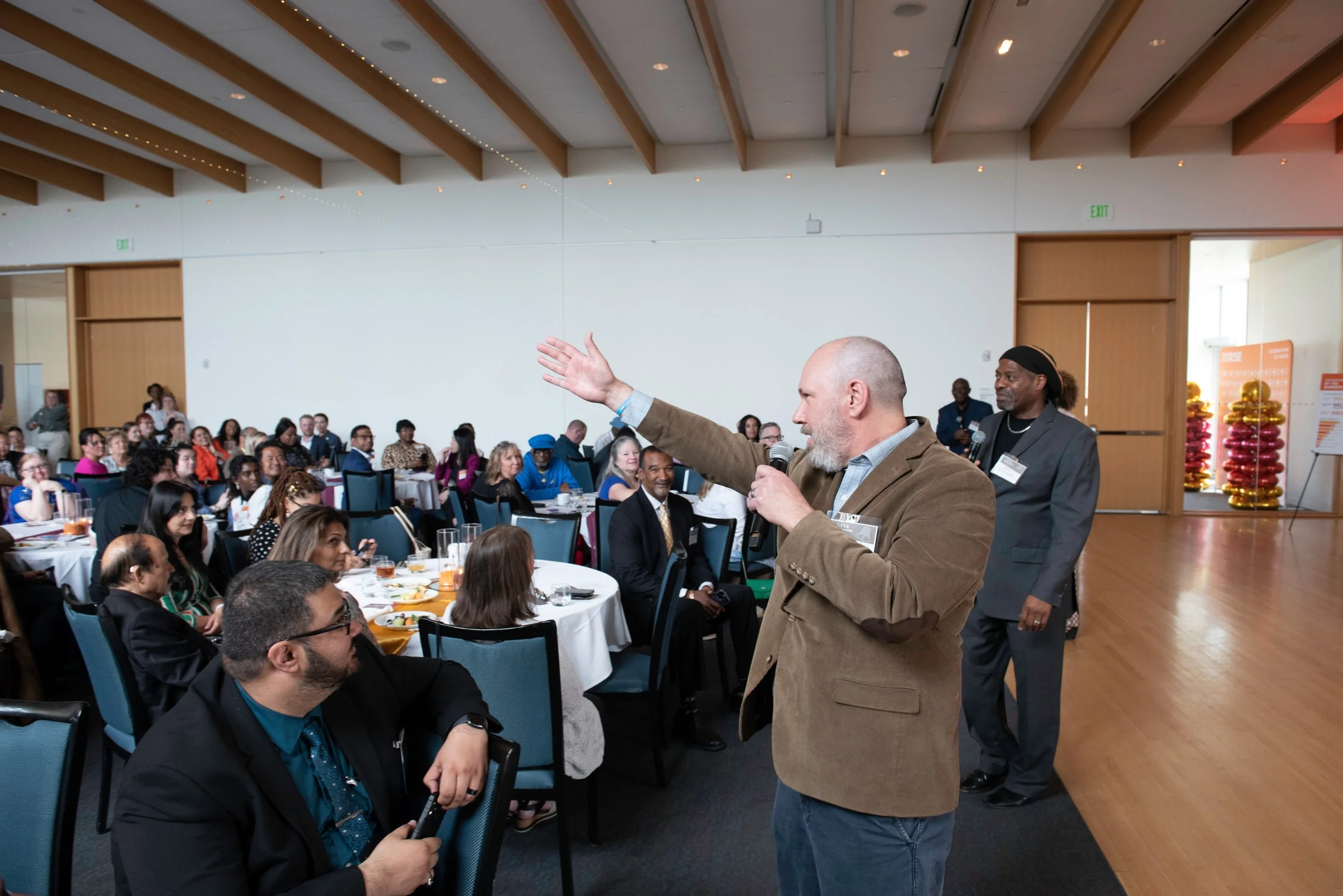 A man with a gray beard, wearing a brown blazer and light blue shirt, speaking into a microphone while raising his hand during a conference. Audience members are seated at round tables with food and drinks, listening attentively.