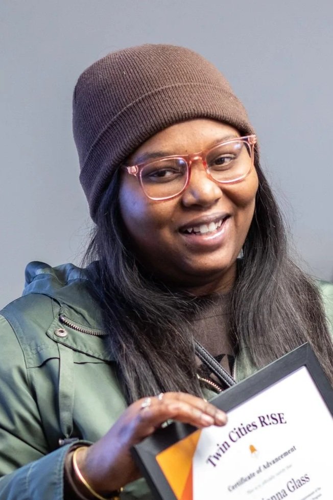A smiling woman wearing glasses and a brown beanie hat, holding a certificate titled "Twin Cities RISE".