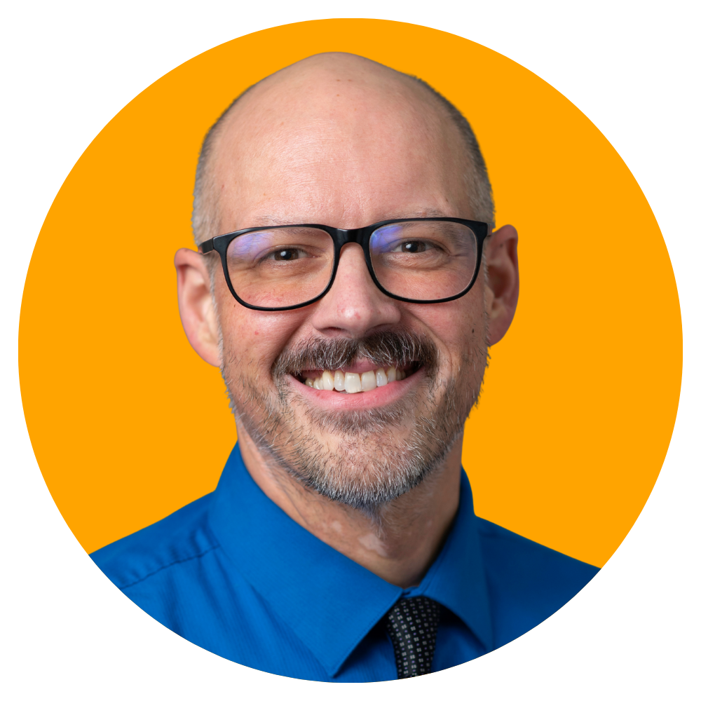 A middle-aged man with glasses, a beard, and a bald head, wearing a blue dress shirt with a patterned tie, smiling against an orange background.