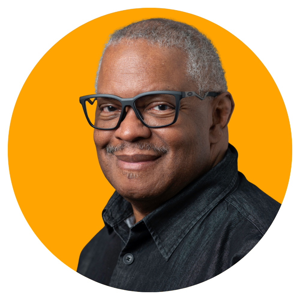A middle-aged man with gray hair, wearing black-rimmed glasses and a black denim shirt, smiling in front of an orange background.