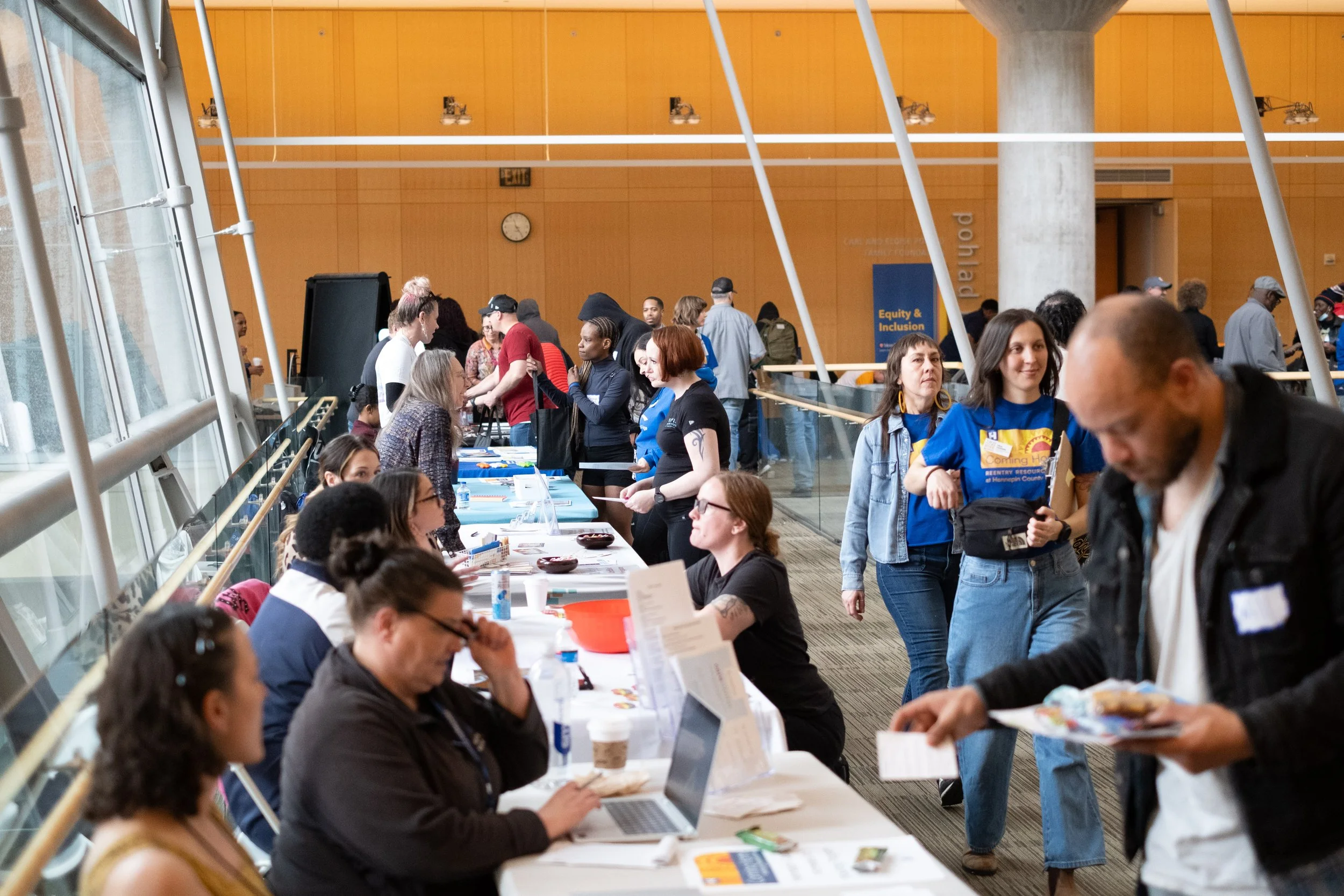 A busy indoor event with people waiting at registration tables and walking through a large hall with orange walls and a clock. Some people are seated and using laptops, while others are standing and talking.