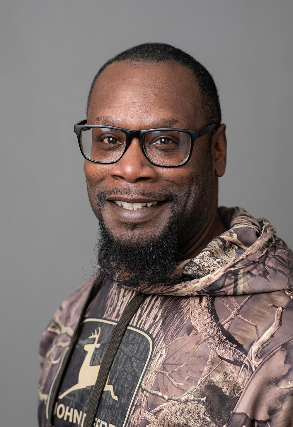 A smiling man wearing glasses and a camouflage hoodie with a John Deere logo, standing against a gray background.