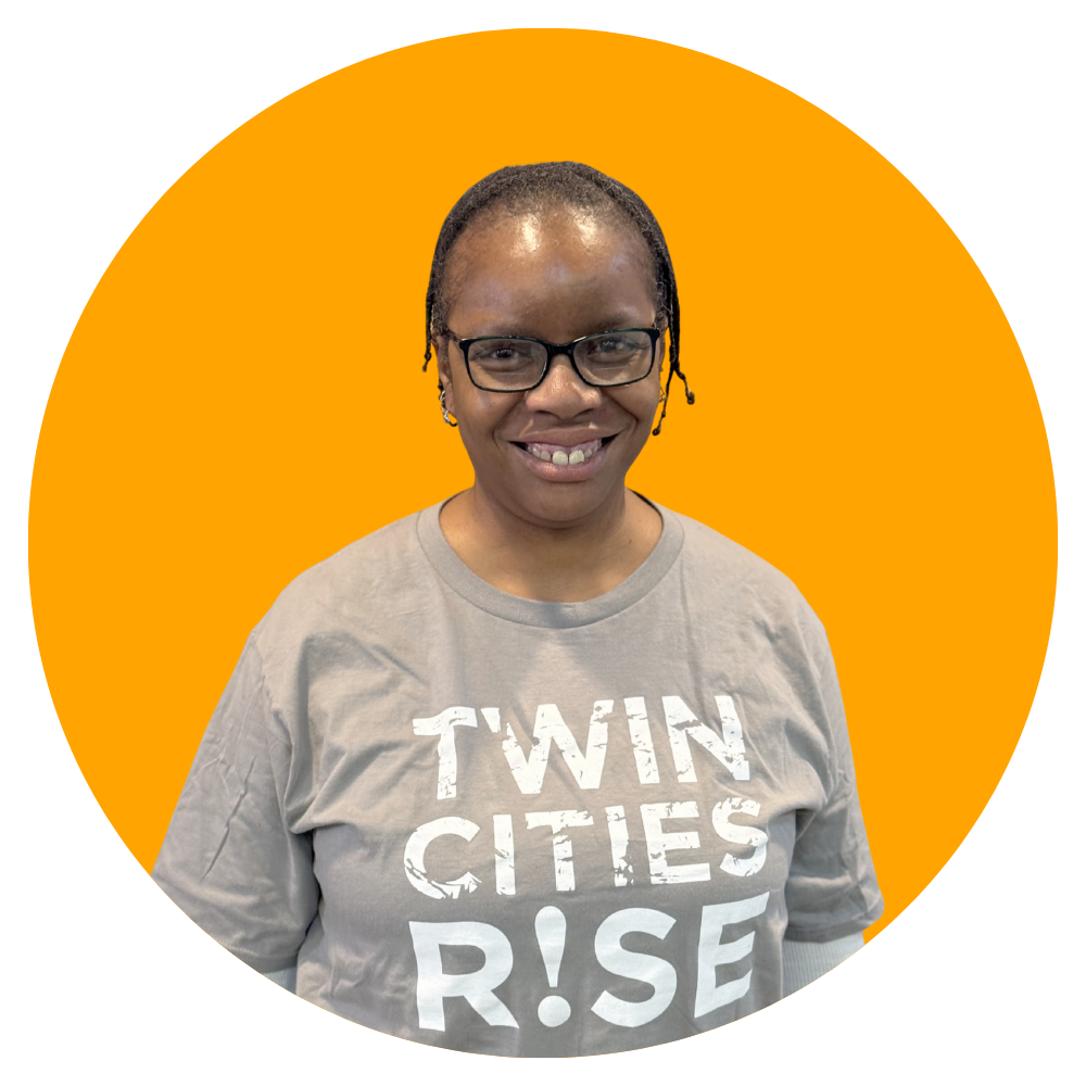A woman with glasses and braided hair smiling, wearing a gray T-shirt that says 'TWIN CITIES RISE'.