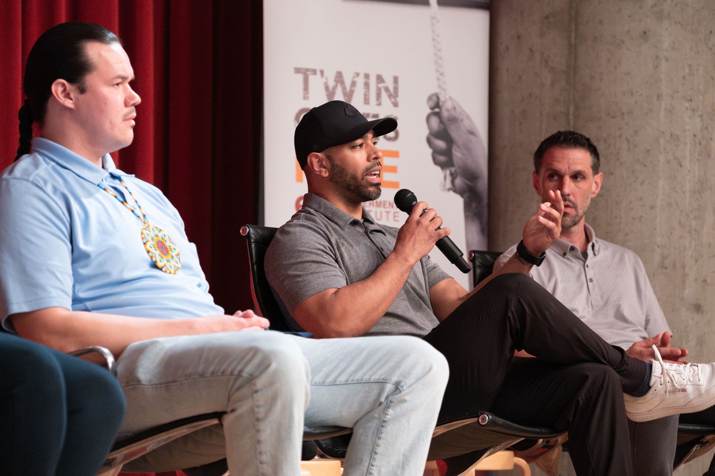 A panel discussion with three men sitting in a row on a stage, one of whom is speaking into a microphone. The background features a red curtain.
