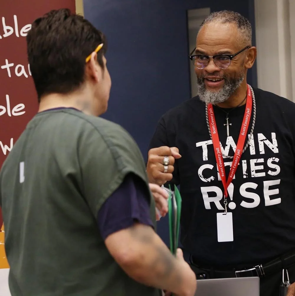 Two men talking at an event, with one wearing a black T-shirt that reads 'TWIN CITIES RISE' and a red lanyard, and the other with glasses and a green jacket, smiling and engaged.