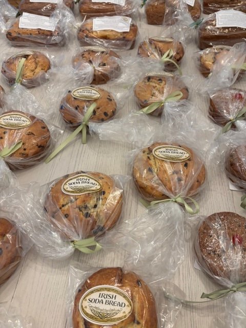 Multiple Irish soda bread loaves wrapped in clear plastic and tied with green ribbons, placed on a table.