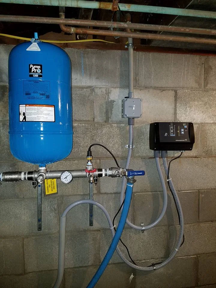 A water pressure booster system with a blue pressure tank, gauges, valves, and control units installed on a concrete wall in a basement.