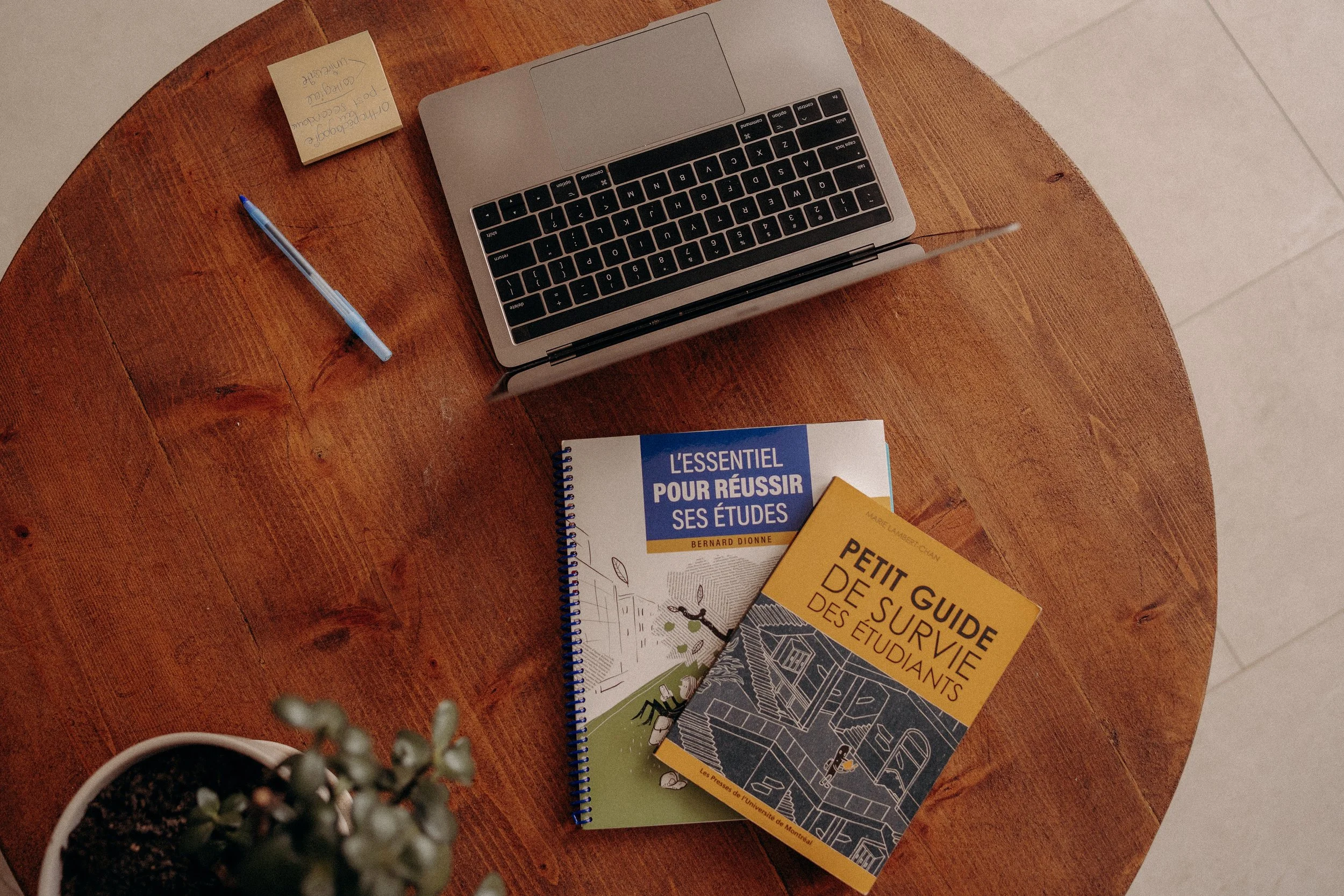 Une table en bois avec un ordinateur portable, des livres, un stylo bleu, une petite note autocollante, et une plante en pot visible sur le coin inférieur gauche.