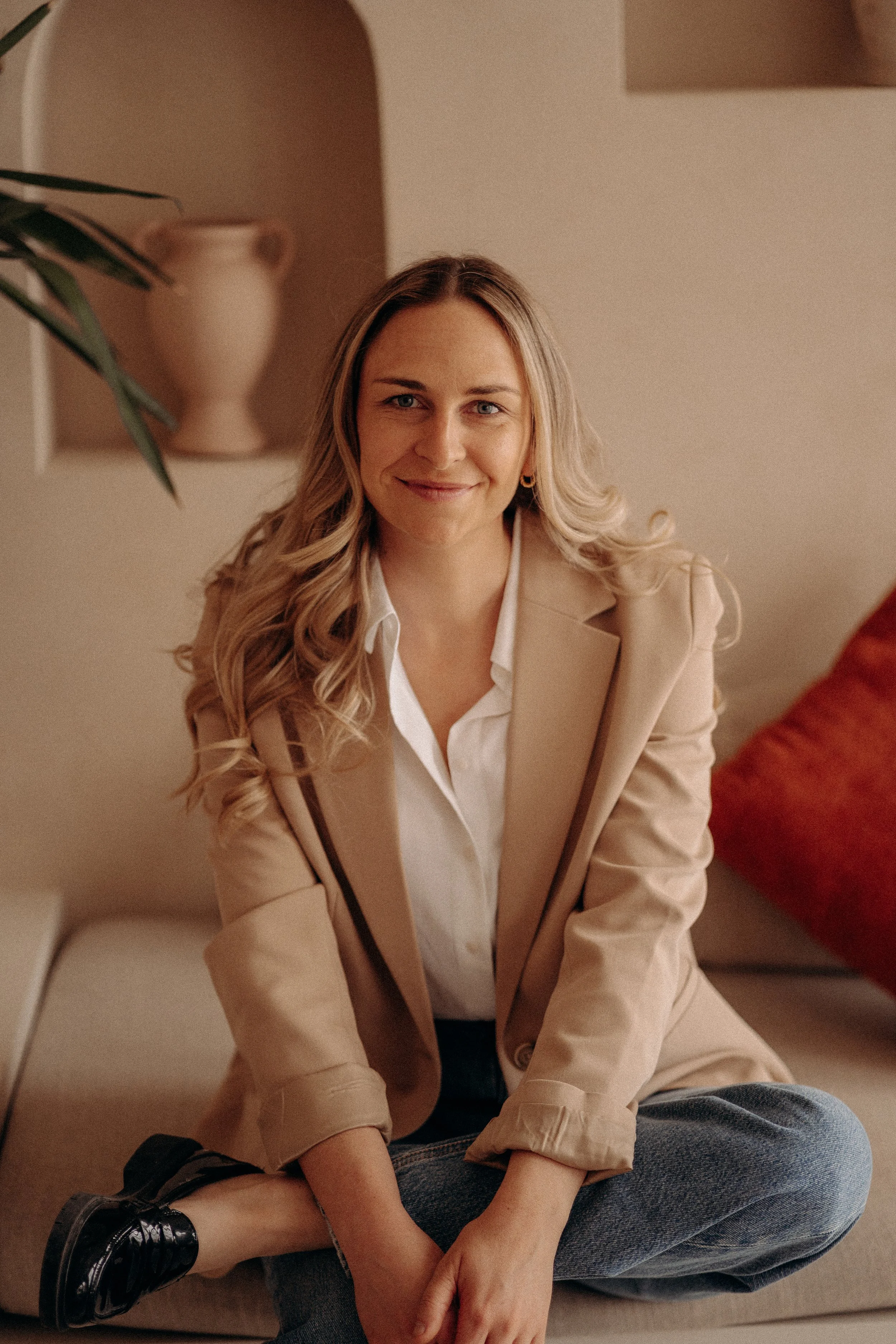 Une femme blonde souriante assise sur un canapé beige, portant une veste beige, une chemise blanche, un jean et des chaussures noires brillantes, avec un vase beige et un mur clair en arrière-plan.