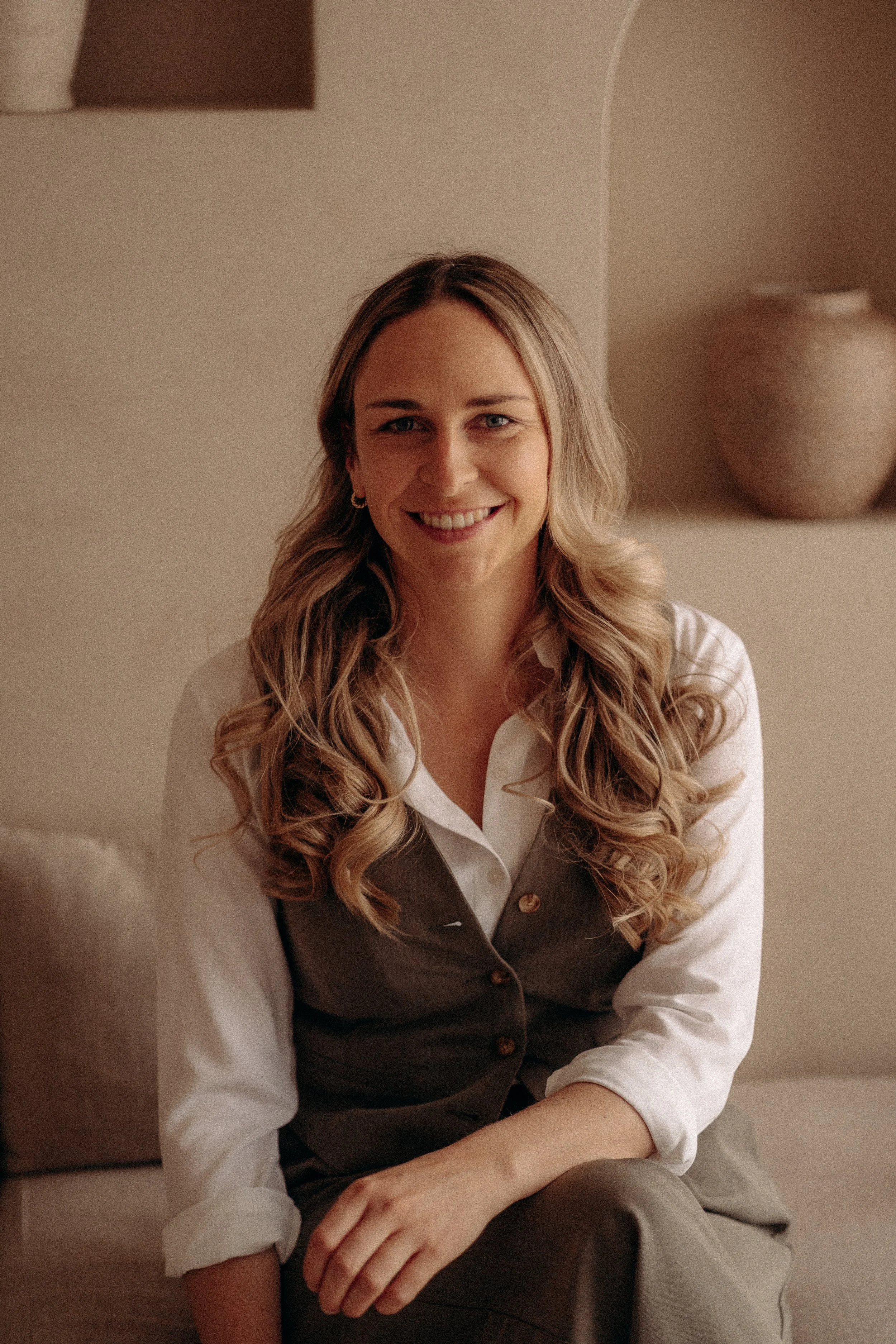 Jeune femme aux cheveux blonds bouclés, souriante, portant une chemise blanche et un gilet marron clair, assise près d'un mur beige avec une ampoule suspendue au plafond.