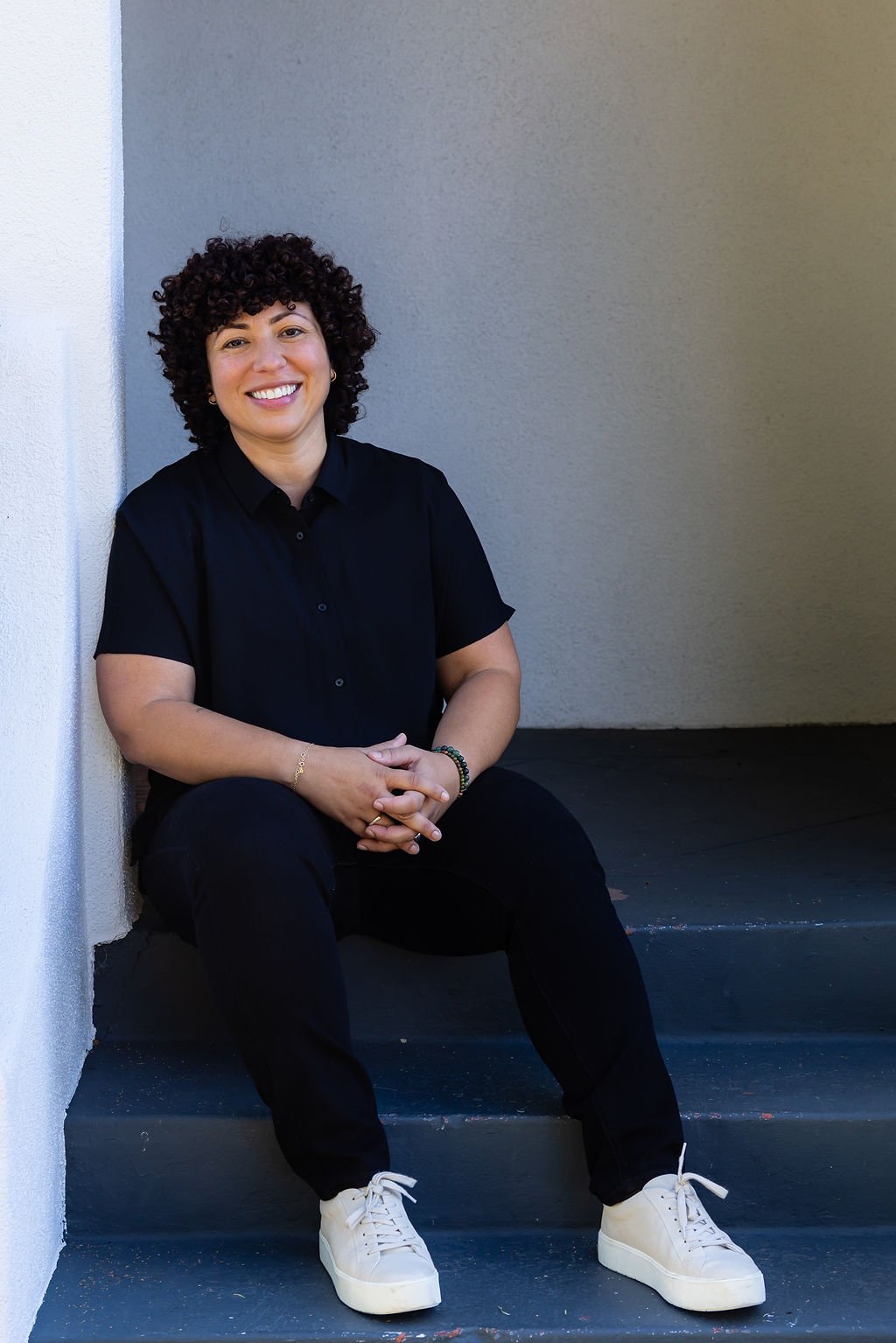 A woman with curly hair sitting on dark stairs, smiling, wearing a black shirt and pants with white sneakers, against a two-tone wall background.