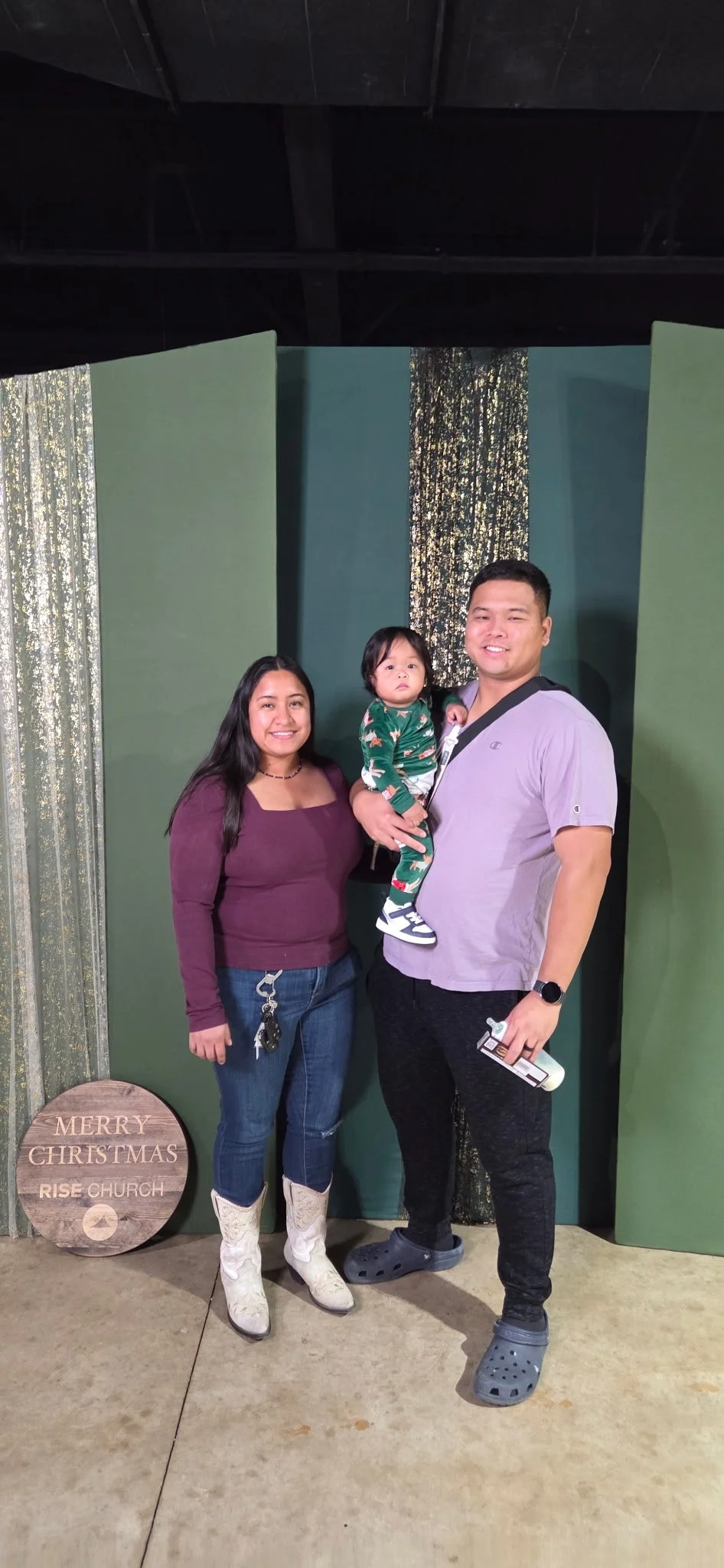 A family of three standing in front of a decorative Christmas backdrop, with a woman and man smiling and a little girl in Christmas pajamas, wearing cowboy boots and Crocs.