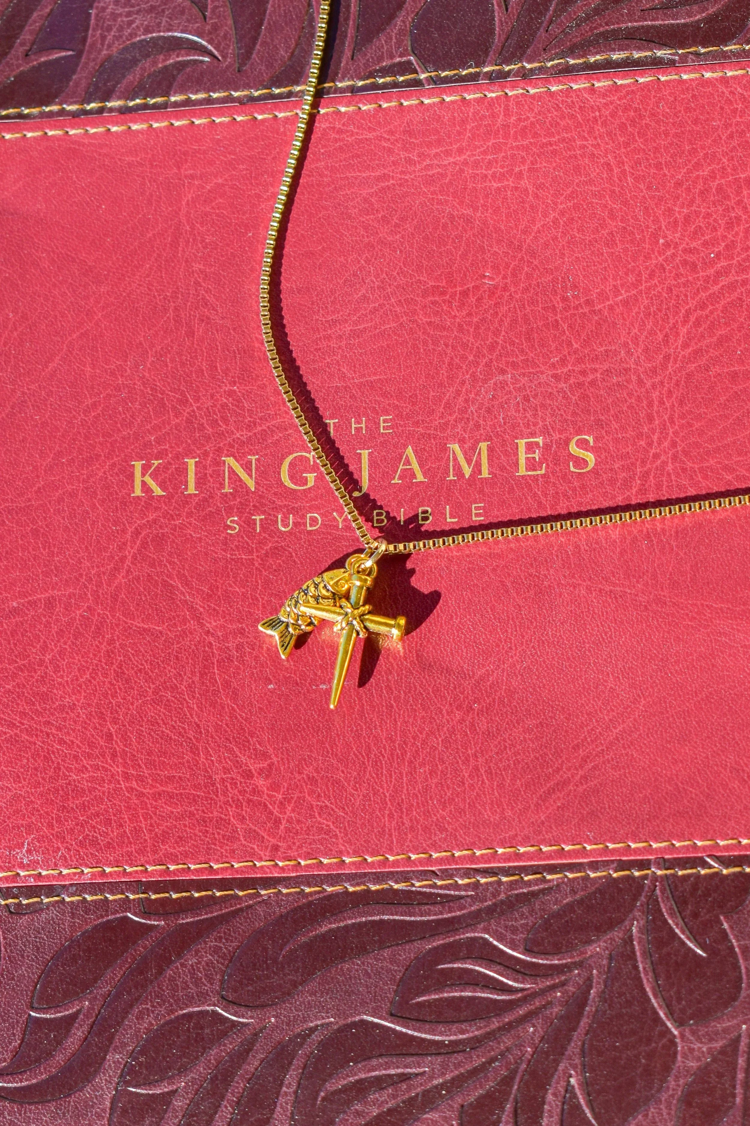 A gold necklace with a pendant of a bee, sword, and shield, resting on a red leather-bound book titled 'The King James Study Bible'.