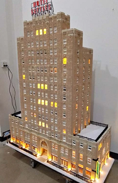 Miniature model of the Hotel Settles building with lit windows and a sign on top, placed indoors on a table.