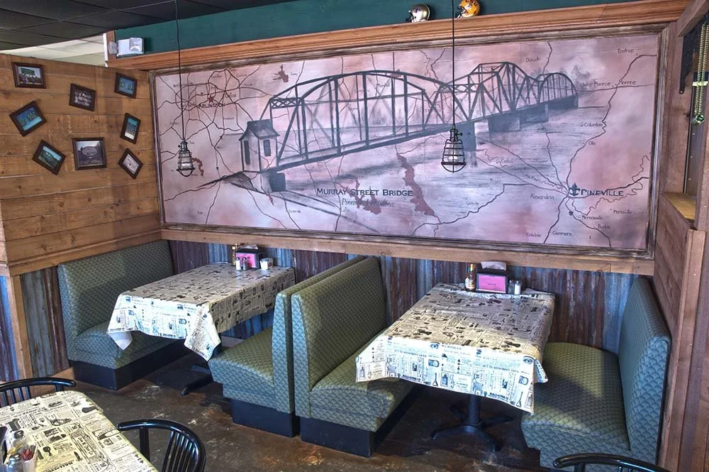 Interior of a restaurant with green booth seating, tables covered with newspaper-patterned tablecloths, and a large mural of a bridge and map of towns on the wall.