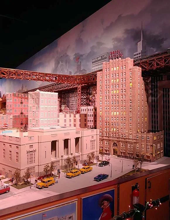 Miniature model of a cityscape with tall buildings, a bridge, and a street with parked retro cars.