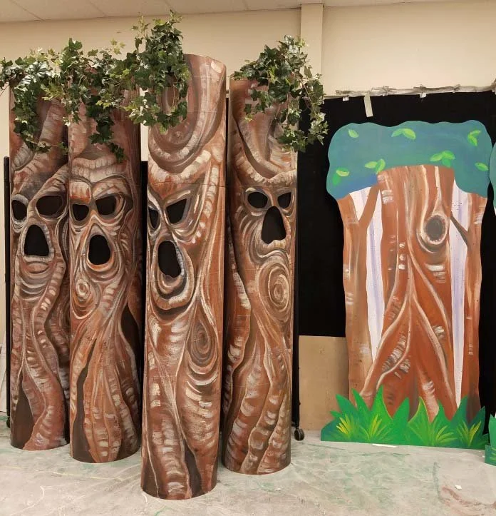Four wooden tree trunk props with facial features, topped with artificial green foliage, next to a painted cardboard cutout of a tree with green leaves and grass at the bottom, in a setting resembling a forest or stage decor.