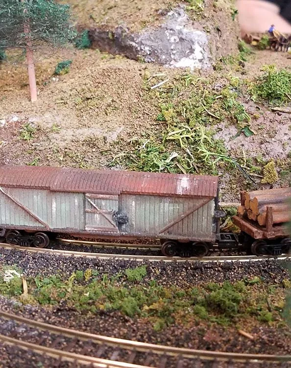 A miniature train with cargo cars on a model railroad track, set in a landscaped outdoor scene.