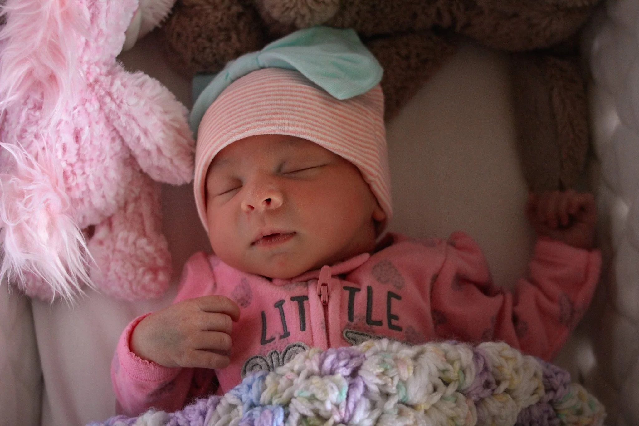 A sleeping baby girl wearing a pink and white striped hat, dressed in pink, lying on a rainbow-colored blanket with a pink fluffy stuffed animal nearby.
