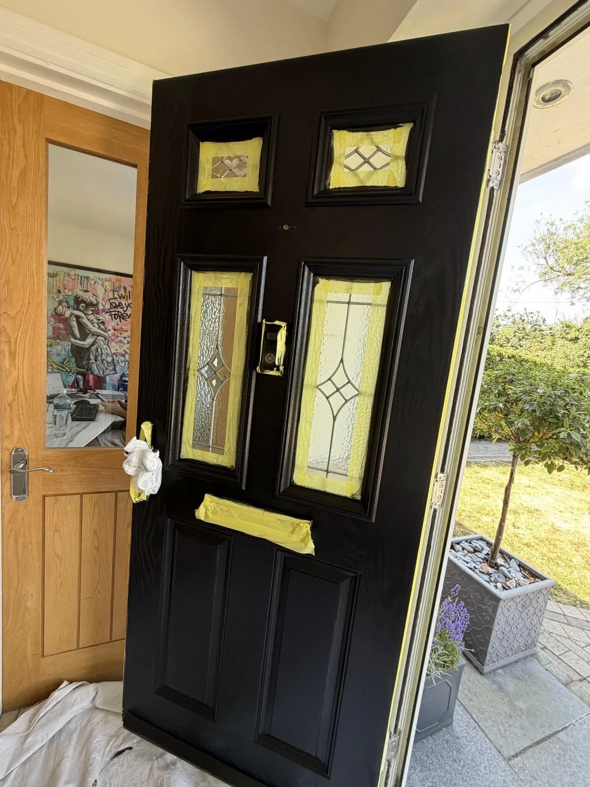Black front door with decorative glass panels, surrounded by yellow painter's tape for painting or refinishing, with a view to the outside showing a tree and garden.