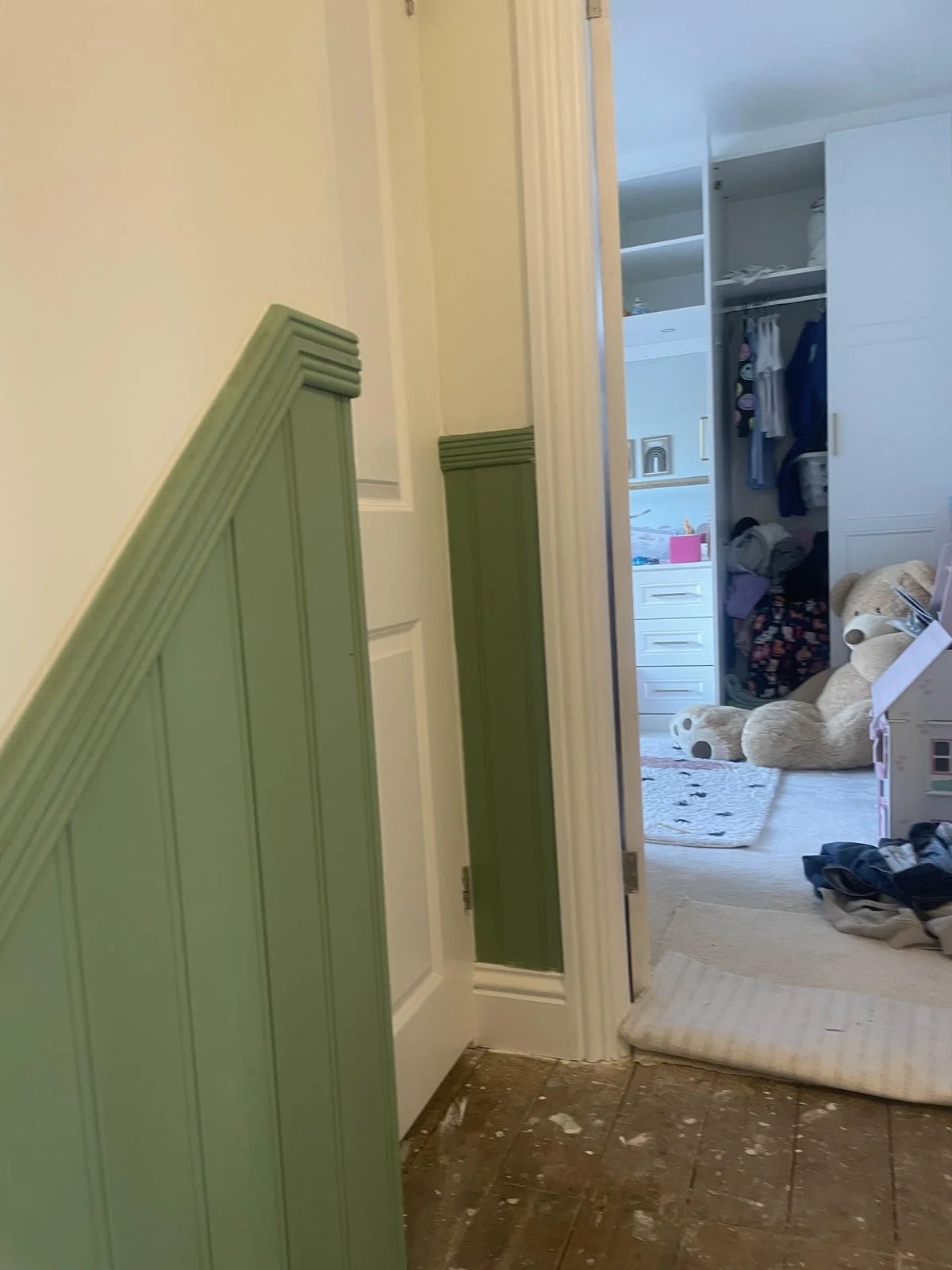 View of a doorway leading into a child's bedroom with a green-wainscoted wall and a carpeted floor. The room contains a large teddy bear, a white dresser, a closet with colorful clothes, and a small playhouse.