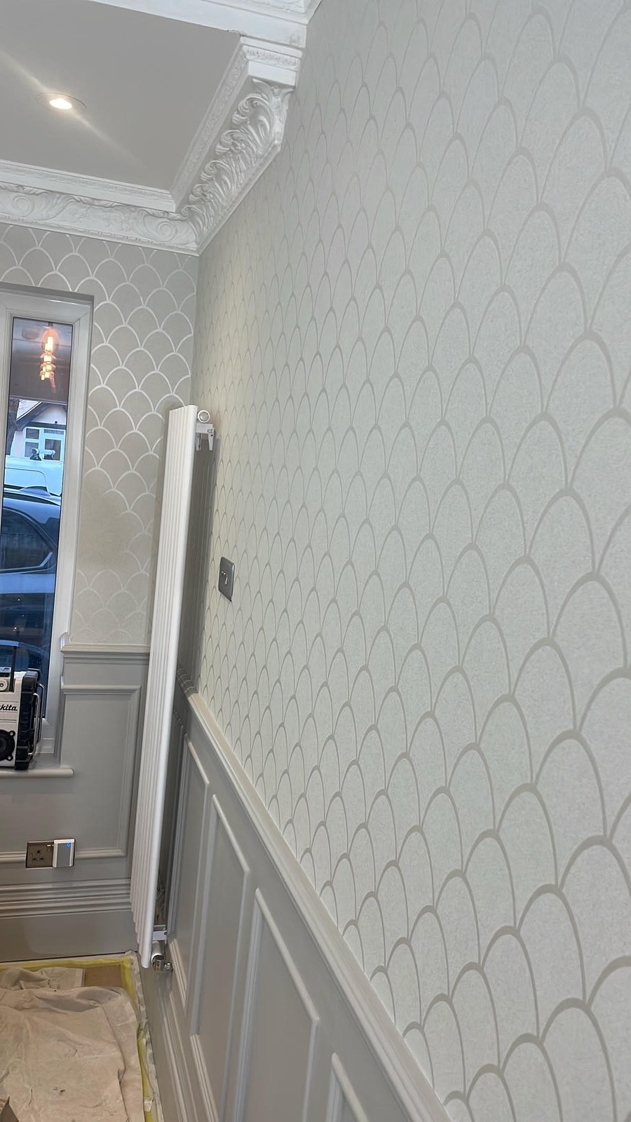 Interior wall with patterned wallpaper, corner view with decorative molding, window with a view of parked cars outside, radiator, electrical outlet, and part of a protective covering on the floor.