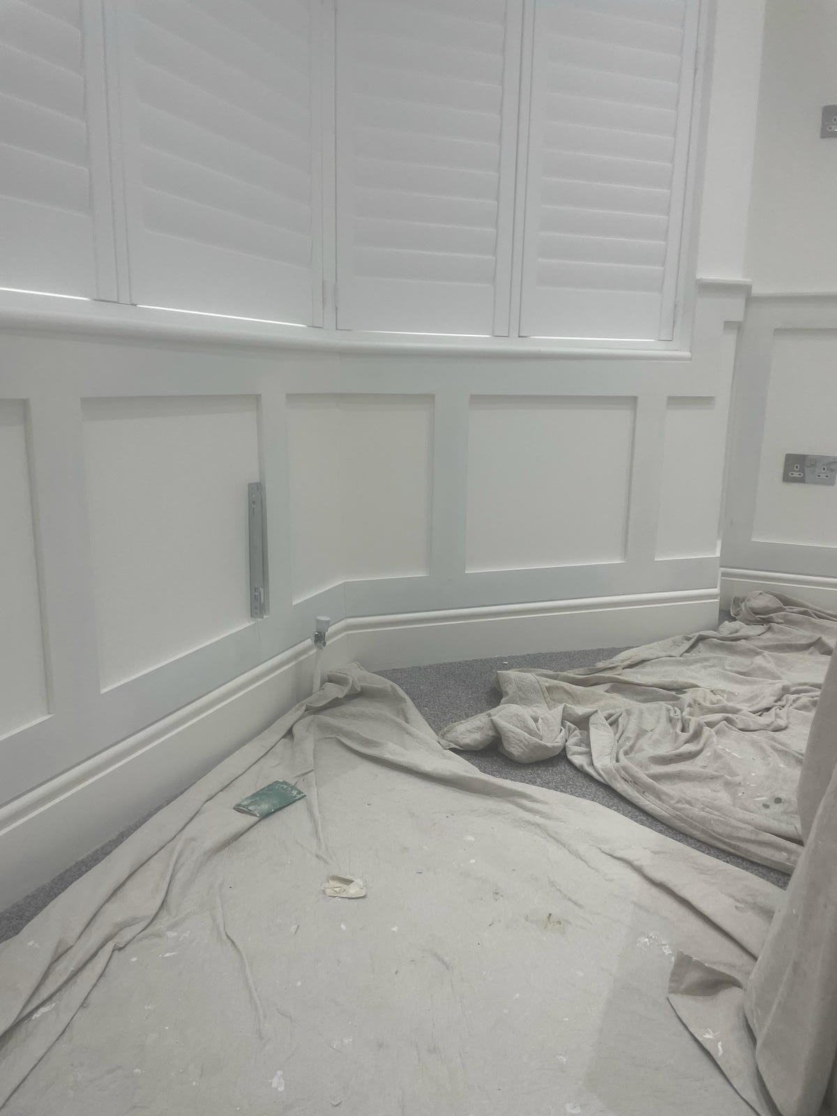 Room under renovation with white wall paneling and shutters, with drop cloths on the floor and construction tools.