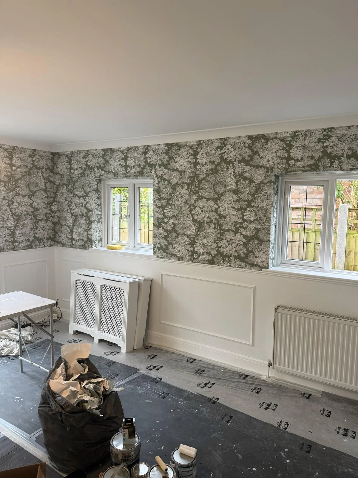 Room undergoing renovation with wallpapered walls, two small windows, and painting supplies on the floor.