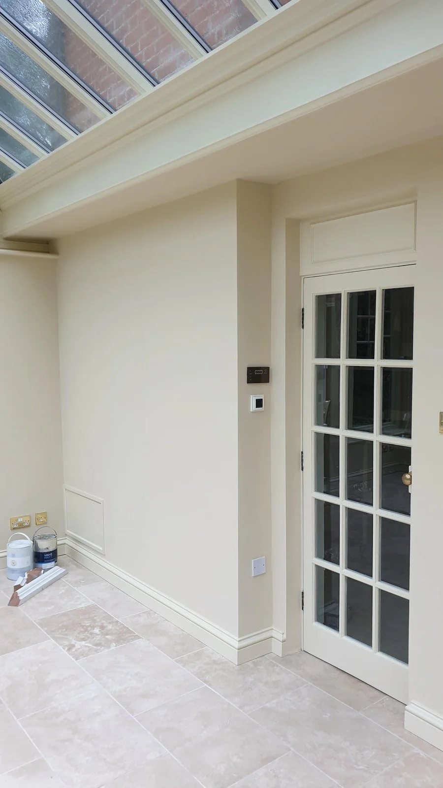 Interior corner of a room with cream-colored walls, large beige tiled floor, a white paneled French door, and a windowed ceiling with wooden framing.