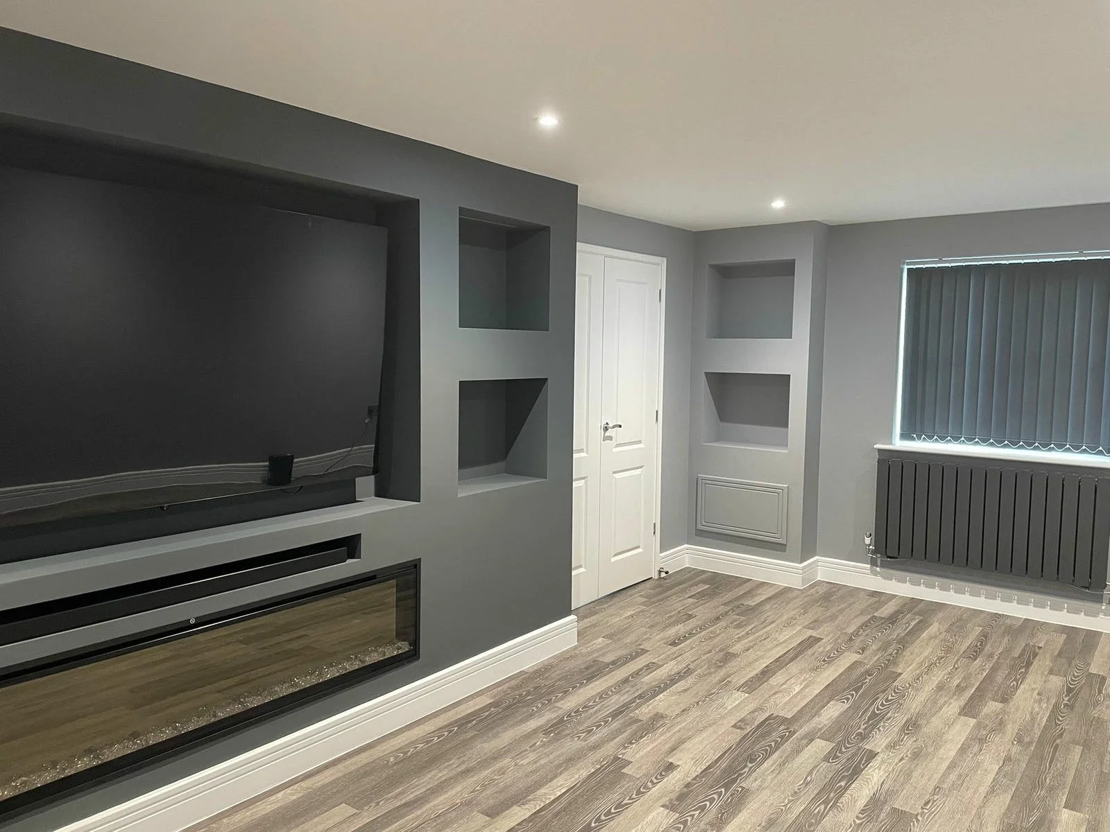 A modern living room with gray walls, wood flooring, a white door, built-in shelves, a large window with vertical blinds, and a wall-mounted electric fireplace beneath a TV.