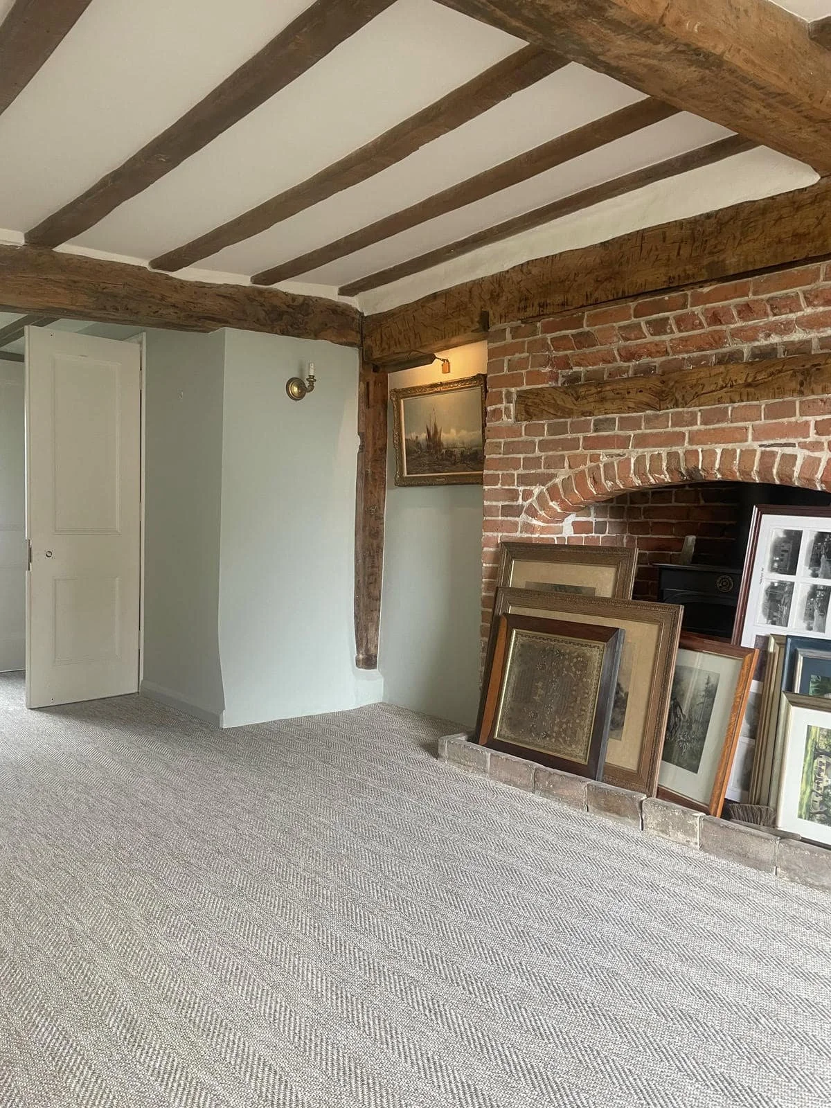 Interior room with exposed wooden beams on a white ceiling, a brick fireplace with framed artwork leaning against it, and a light-colored carpeted floor.