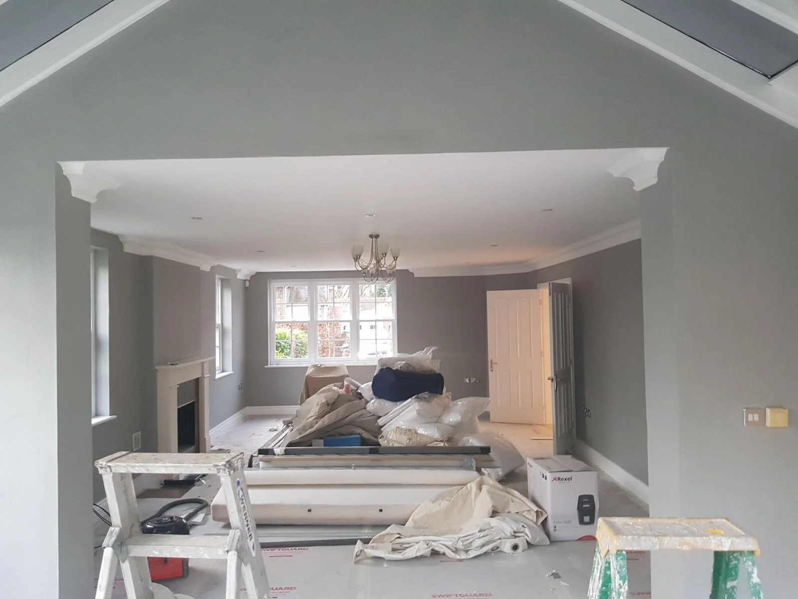 Living room under renovation with construction tools and supplies, including ladders, debris, and packages, in front of a gray-painted wall.