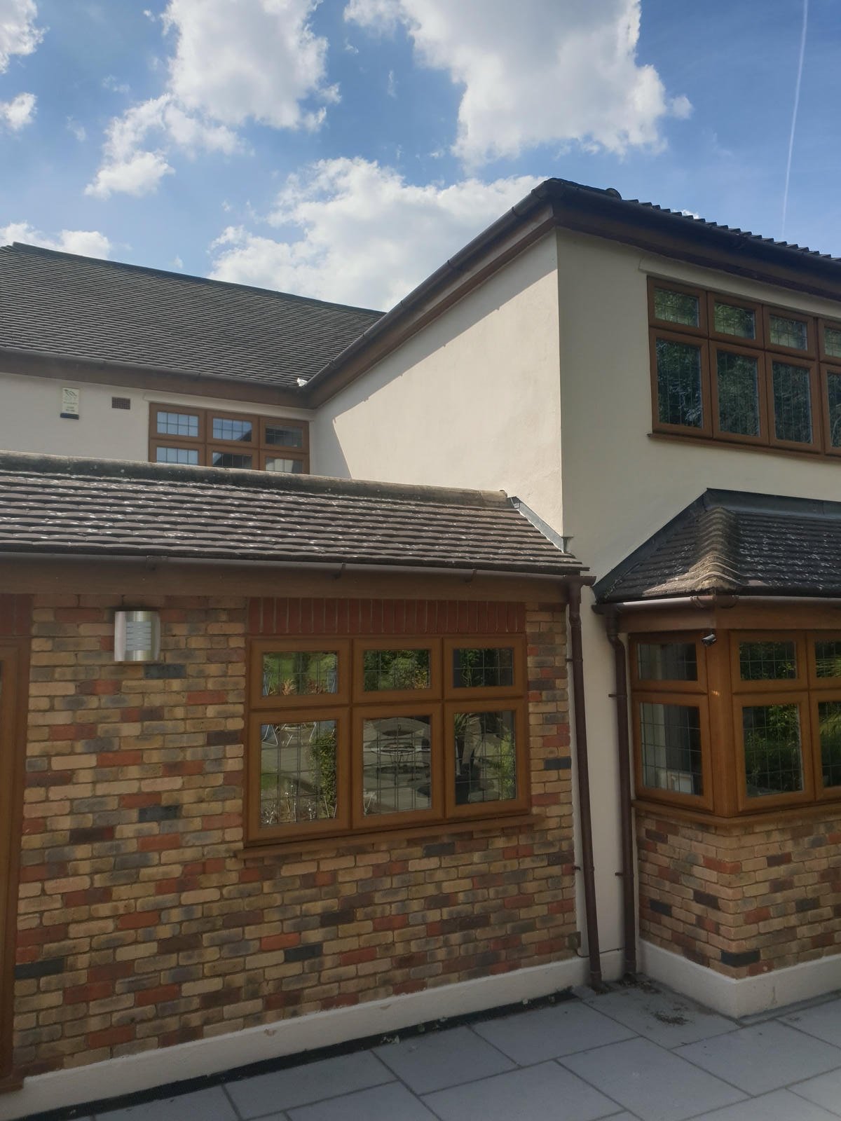 Part of a house with brick and white walls, brown window frames, tiled roof, and a clear blue sky with clouds.