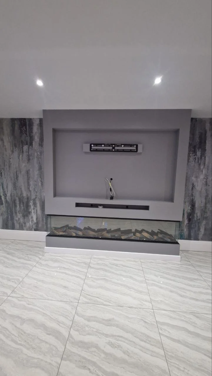 Unfinished modern fireplace with a gray frame, a glass panel filled with decorative stones, and a mounted TV bracket with wires hanging.
