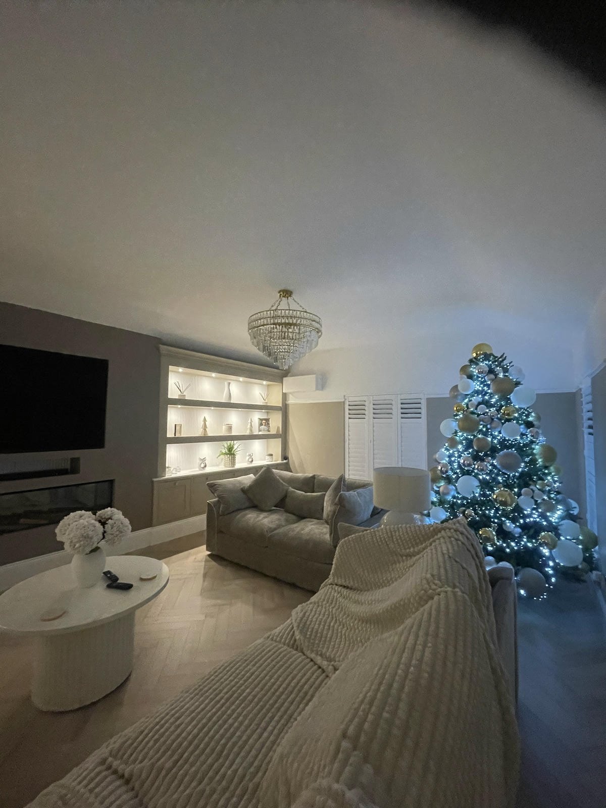 Living room decorated for Christmas with a lit Christmas tree, a chandelier, a wall-mounted TV, a built-in display shelf with decorations, a beige couch with pillows, a coffee table with flowers, and a blanket draped over another piece of furniture.